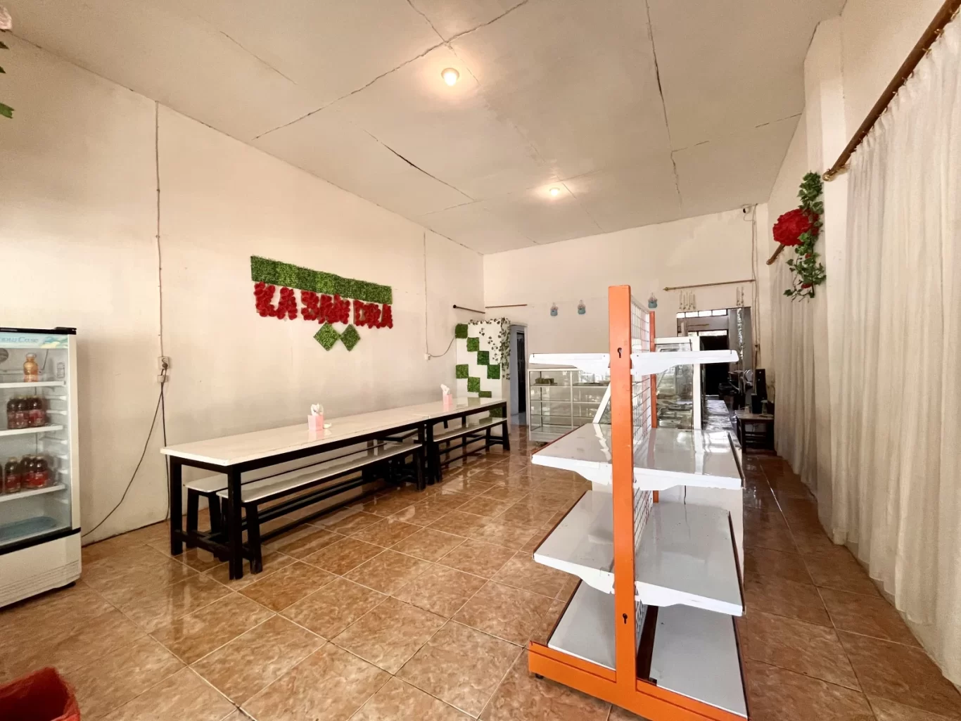 The interior of Depot Kassandra shows long white tables with benches, a display fridge, and empty orange-and-white shelves on a tiled floor.