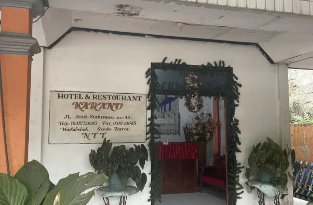The entrance of Hotel Karanu features a white tiled floor, potted green plants, and a sign for "Hotel & Restaurant Karanu" on a white wall, leading to a decorated doorway.