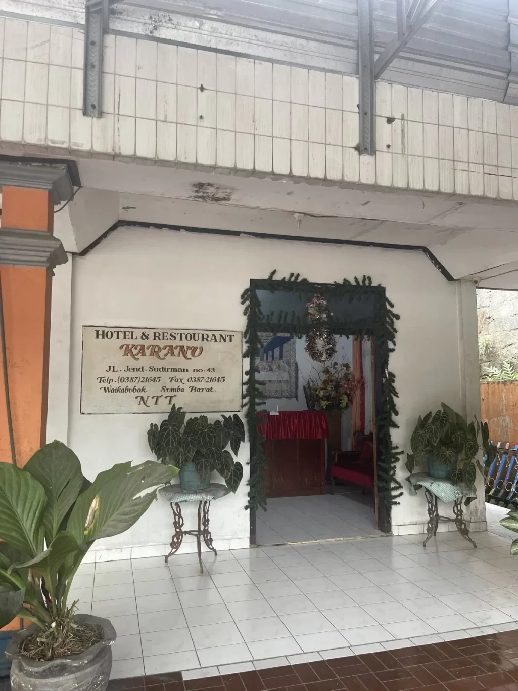 The entrance of Hotel Karanu features a white tiled floor, potted green plants, and a sign for "Hotel & Restaurant Karanu" on a white wall, leading to a decorated doorway.