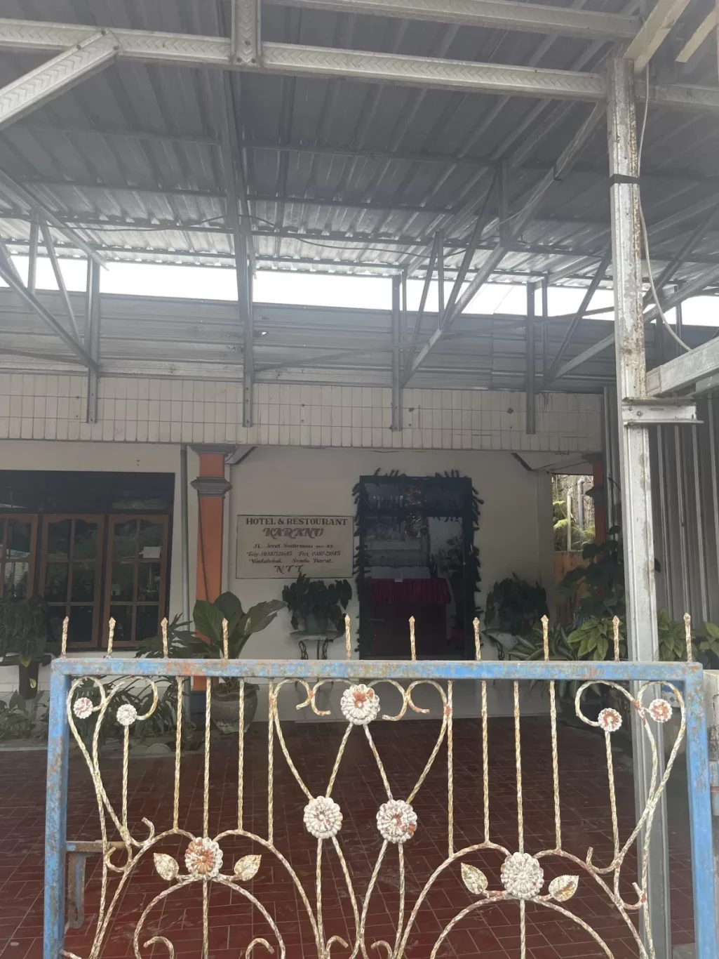 A blue and gold ornate metal gate stands partially open, revealing a white "Hotel & Restaurant Karanu" sign, green plants, and a metal roof structure.