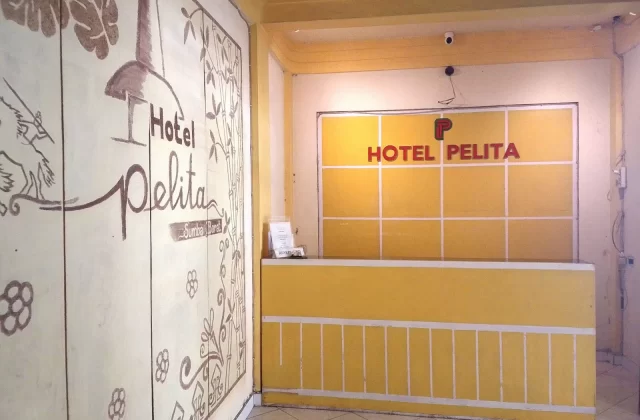 The reception area of Hotel Pelita features a yellow desk and a wall with "HOTEL PELITA" in red, next to a cream wall with brown traditional Sumba illustrations.