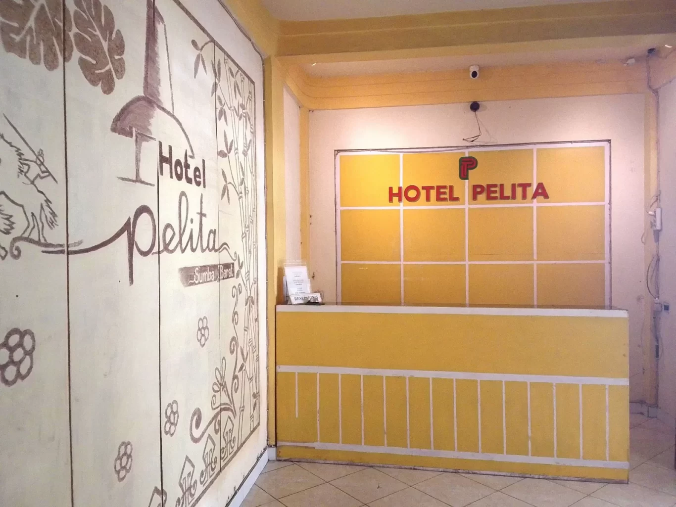 The reception area of Hotel Pelita features a yellow desk and a wall with "HOTEL PELITA" in red, next to a cream wall with brown traditional Sumba illustrations.