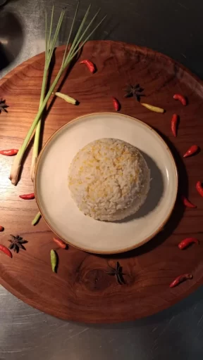 A white plate with rice is on a dark wooden board, surrounded by green lemongrass, red and yellow chili peppers, and star anise.