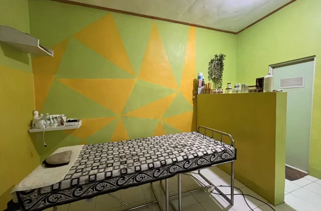 A spa treatment room features a massage bed with a patterned cover, lime green and yellow geometric walls, and shelves with various spa products.