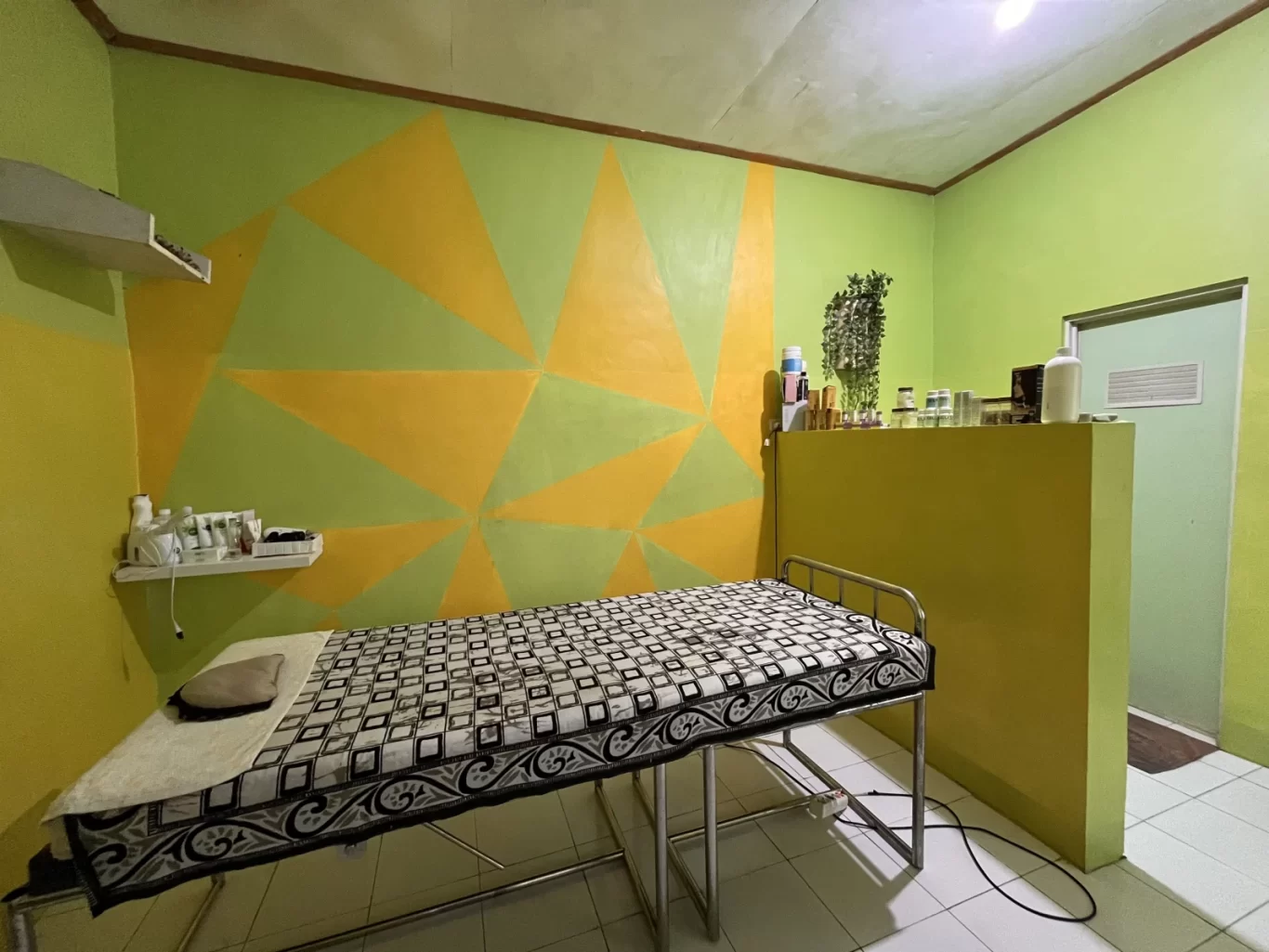 A spa treatment room features a massage bed with a patterned cover, lime green and yellow geometric walls, and shelves with various spa products.