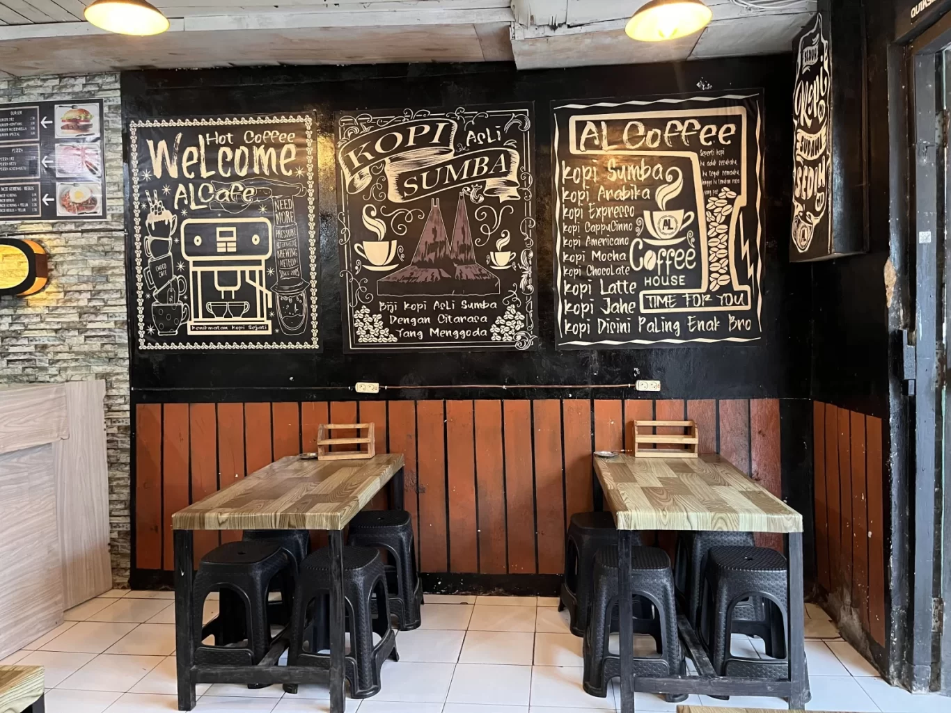 Interior view of AL Cafe in Waikabubak, featuring two light wood tables with black stools against a dark wall adorned with white chalk art and orange wooden paneling.