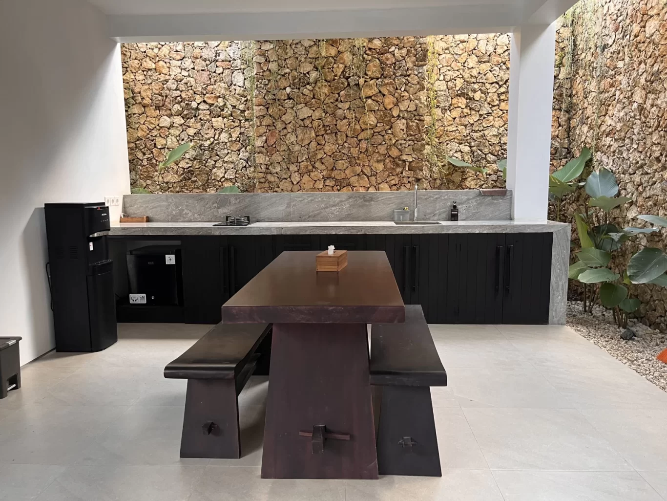 A modern outdoor kitchen and dining area features natural stone walls, a dark wooden table with benches, black cabinets, and a water dispenser.