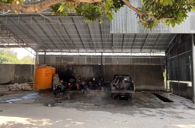 Workers wash a black pickup truck and two motorcycles under a corrugated metal roof at a vehicle wash bay, with a large orange water tank visible.