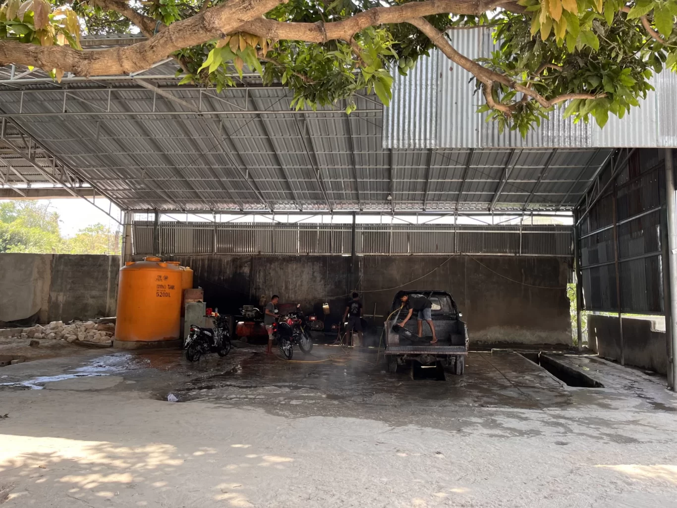 Workers wash a black pickup truck and two motorcycles under a corrugated metal roof at a vehicle wash bay, with a large orange water tank visible.