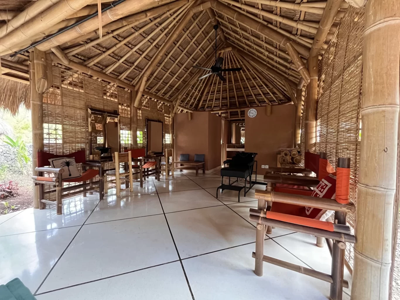 A spacious open-air spa pavilion, constructed from bamboo and thatch, features light tiled floors, bamboo chairs with orange cushions, and mirrors.