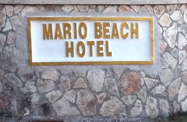 A stone wall features a white rectangular sign with raised gold letters spelling "MARIO BEACH HOTEL" against a backdrop of varied grey and brown stones.