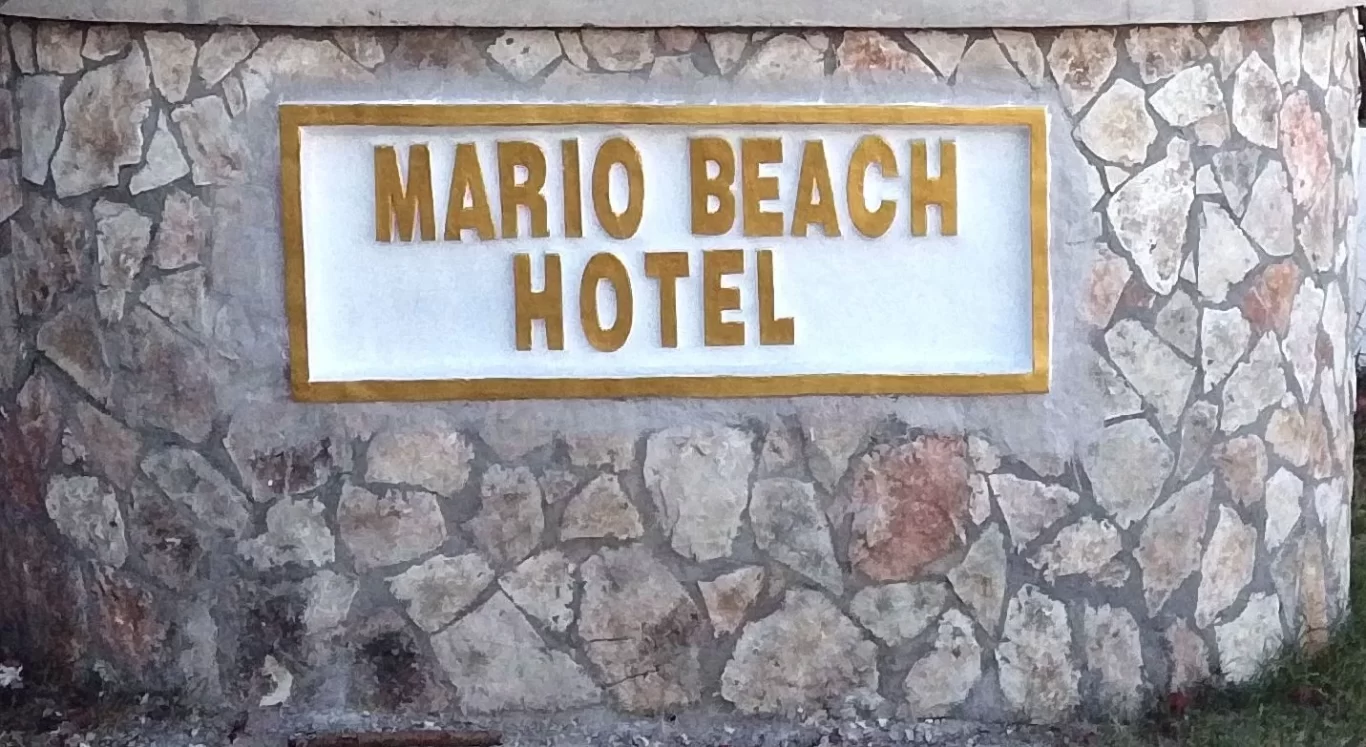 A stone wall features a white rectangular sign with raised gold letters spelling "MARIO BEACH HOTEL" against a backdrop of varied grey and brown stones.
