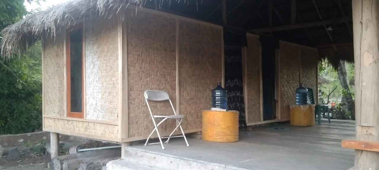 A traditional homestay building with woven bamboo walls and a thatched roof stands on a porch, featuring a folding chair and water bottles, surrounded by green foliage.