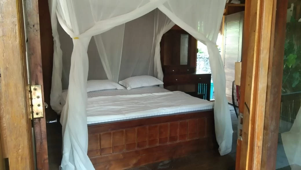 A rustic bedroom features a wooden bed with white bedding and a draped white mosquito net, framed by wooden walls and an open doorway.