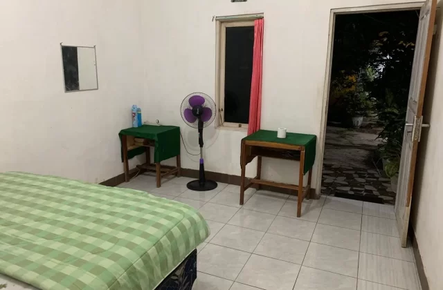 A simple homestay room features a bed with a green checkered blanket, a purple fan, two small tables, and an open door revealing a dark garden.