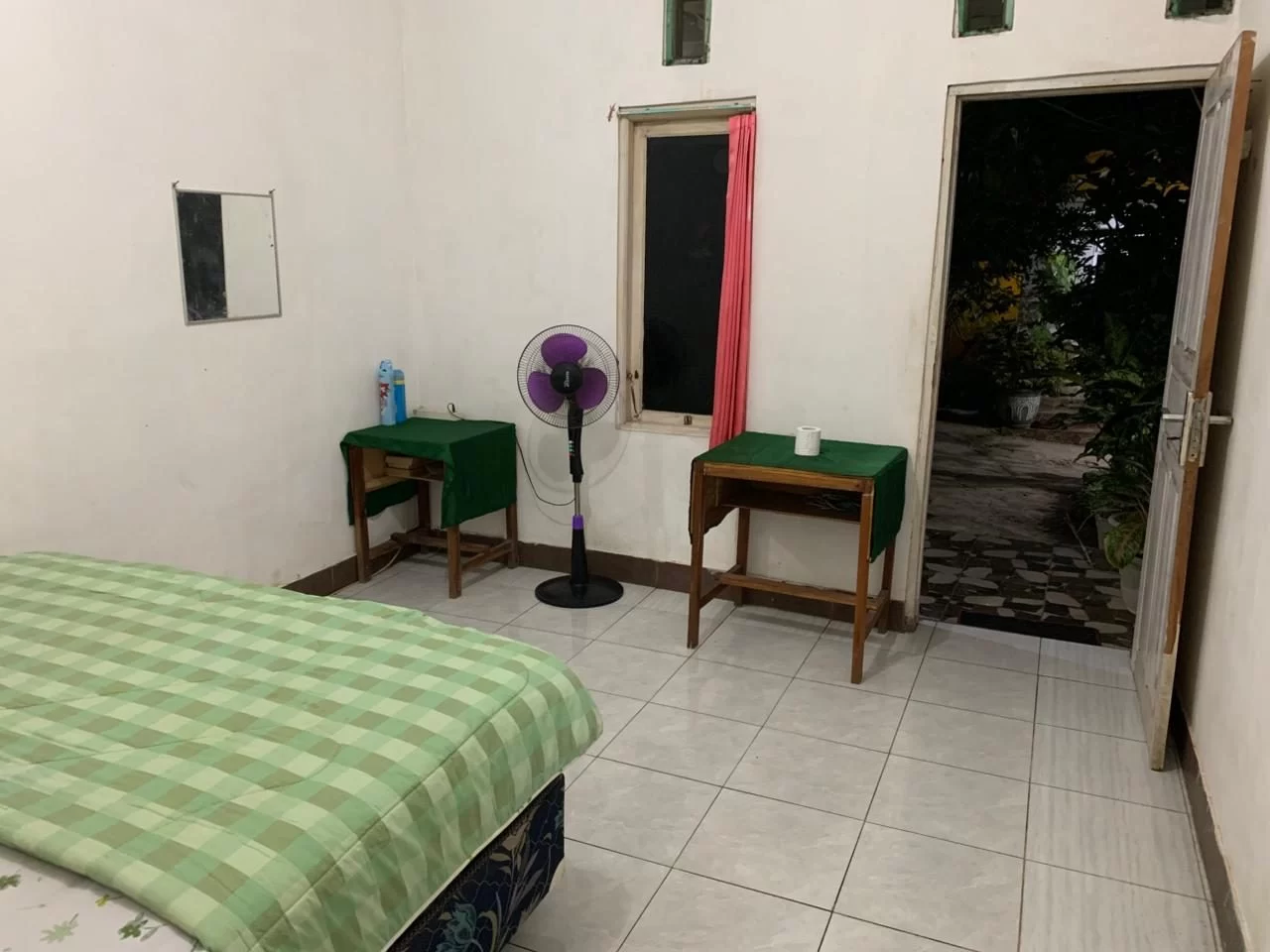 A simple homestay room features a bed with a green checkered blanket, a purple fan, two small tables, and an open door revealing a dark garden.