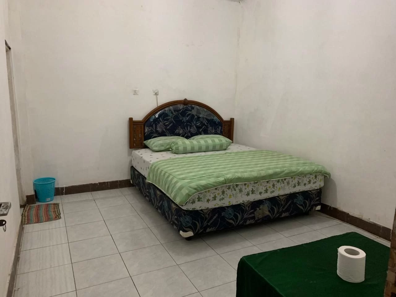 A simple bedroom features a bed with a dark blue patterned base and green striped bedding, white walls, a tiled floor, and a blue trash can.