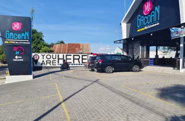 A modern black Mie Gacoan restaurant building and a tall sign stand beside a paved parking lot with cars and a scooter under a clear blue sky.