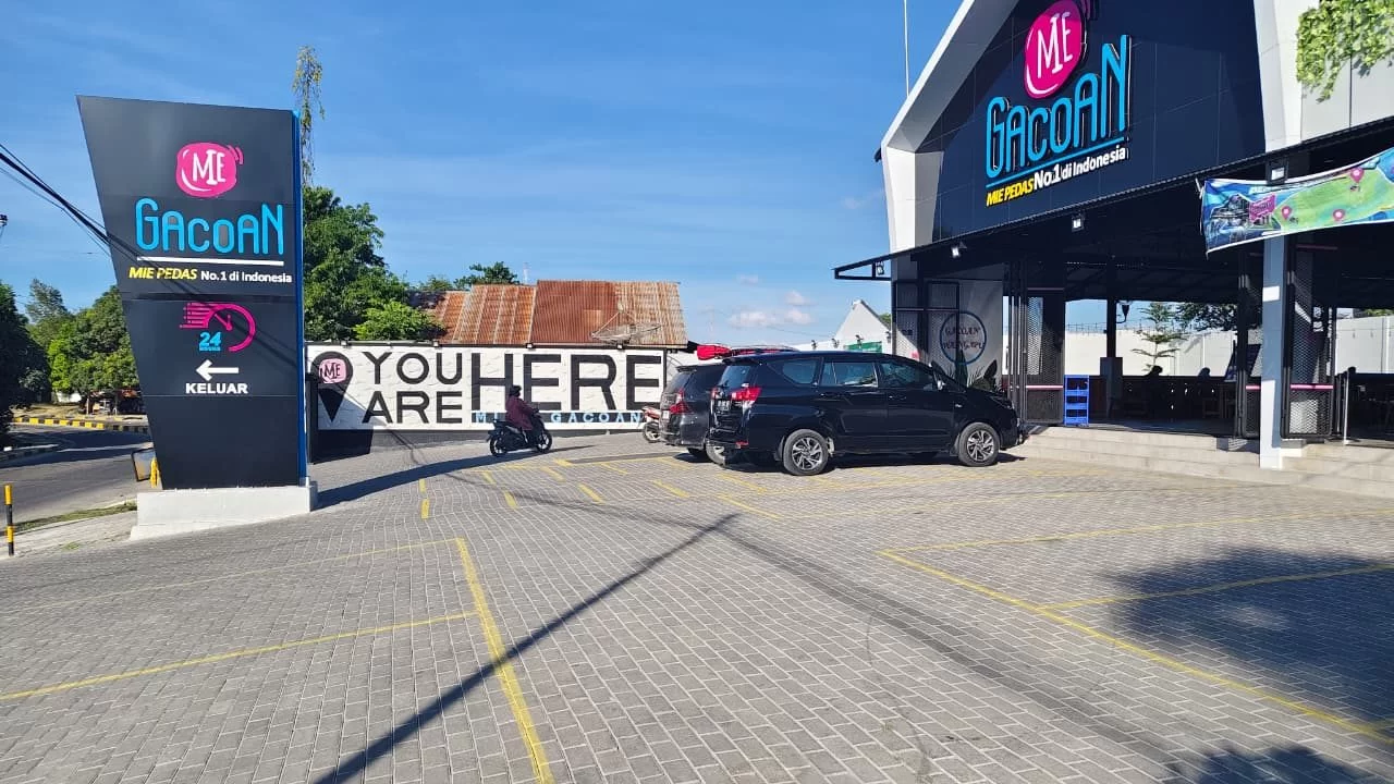 A modern black Mie Gacoan restaurant building and a tall sign stand beside a paved parking lot with cars and a scooter under a clear blue sky.