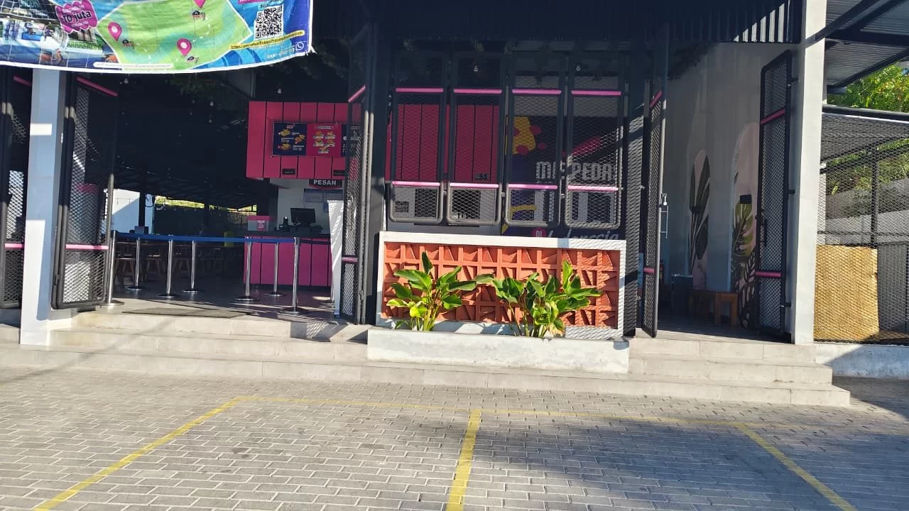 A modern building with pink and black accents, an open entrance, and a banner overhead, viewed from a paved parking lot with yellow lines.