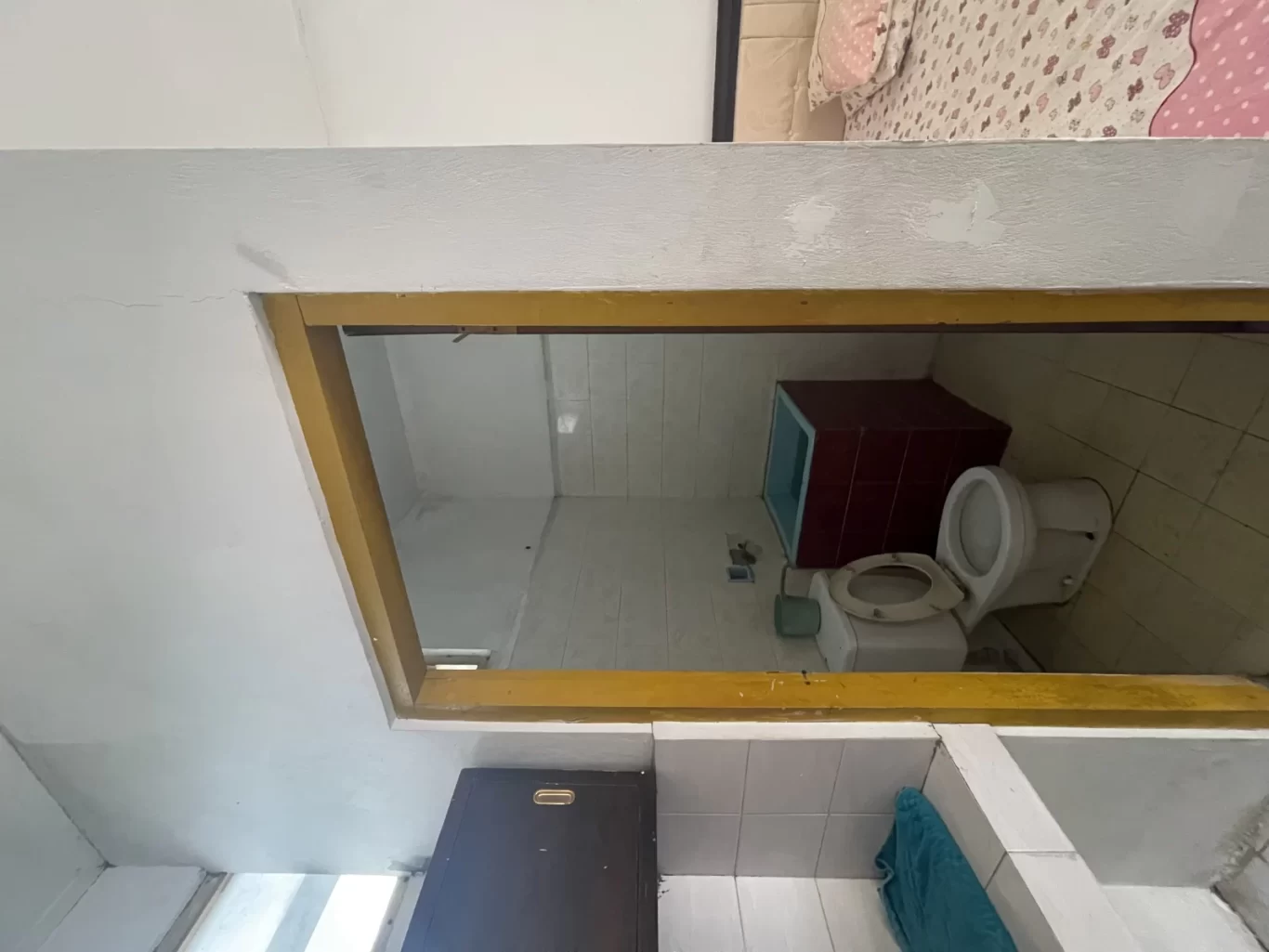 A simple hotel bathroom with white tiled walls, a toilet, and a small red cabinet is seen through a yellow doorway. A bed with a pink duvet is visible above the opening.