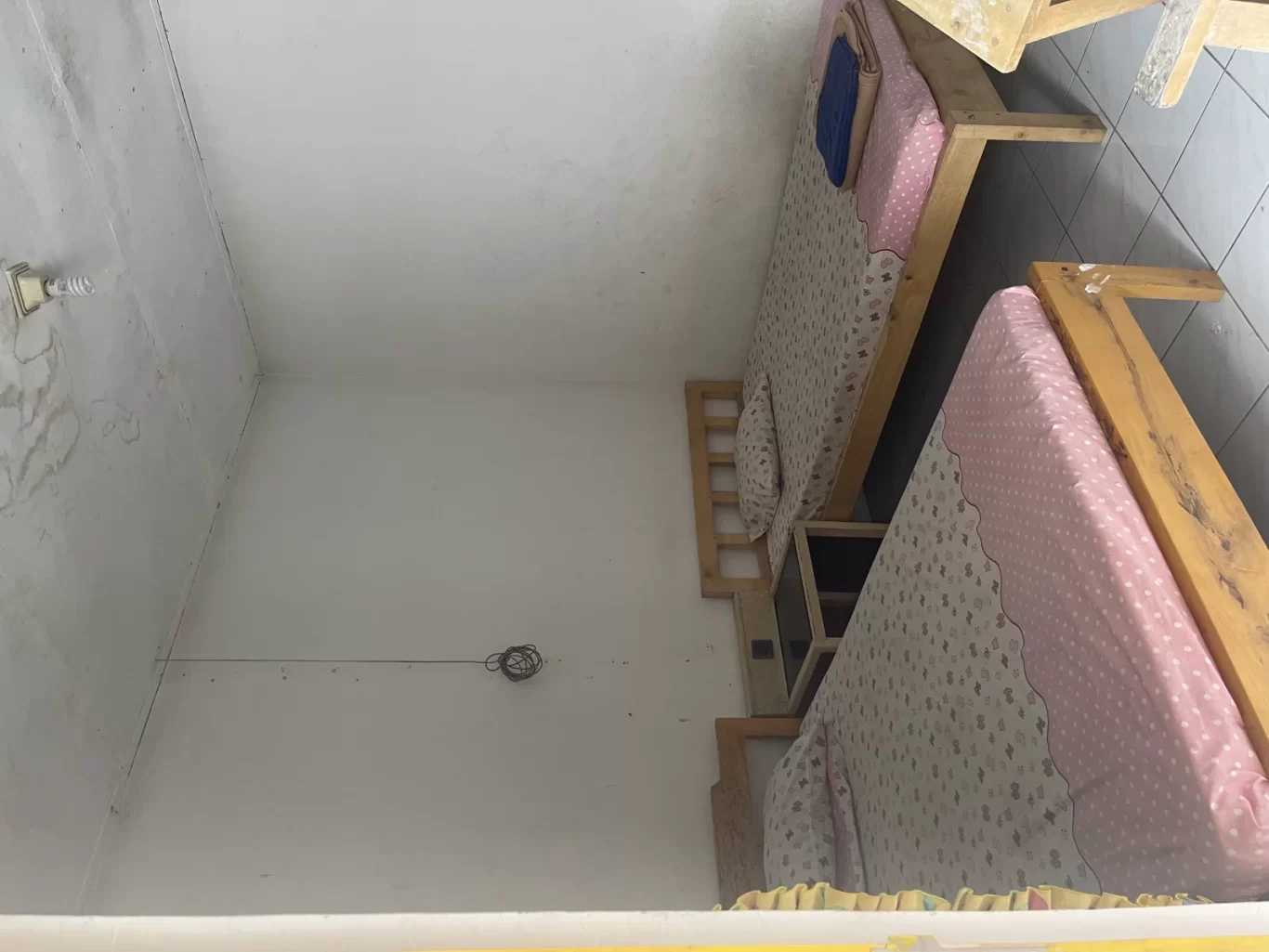 A simple hotel room features two single beds with wooden frames and pink polka dot sheets, a small bedside table, and plain white walls with some discoloration.