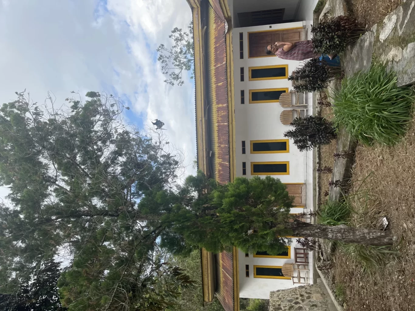 A white hotel building with a rusty roof and yellow-framed windows, surrounded by green trees and dry ground, with a person standing near the entrance under a cloudy sky.