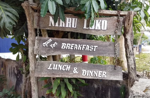 A weathered wooden sign for "Nirru Loko" hangs among green leaves, displaying "Open for Breakfast, Lunch & Dinner" in white text.