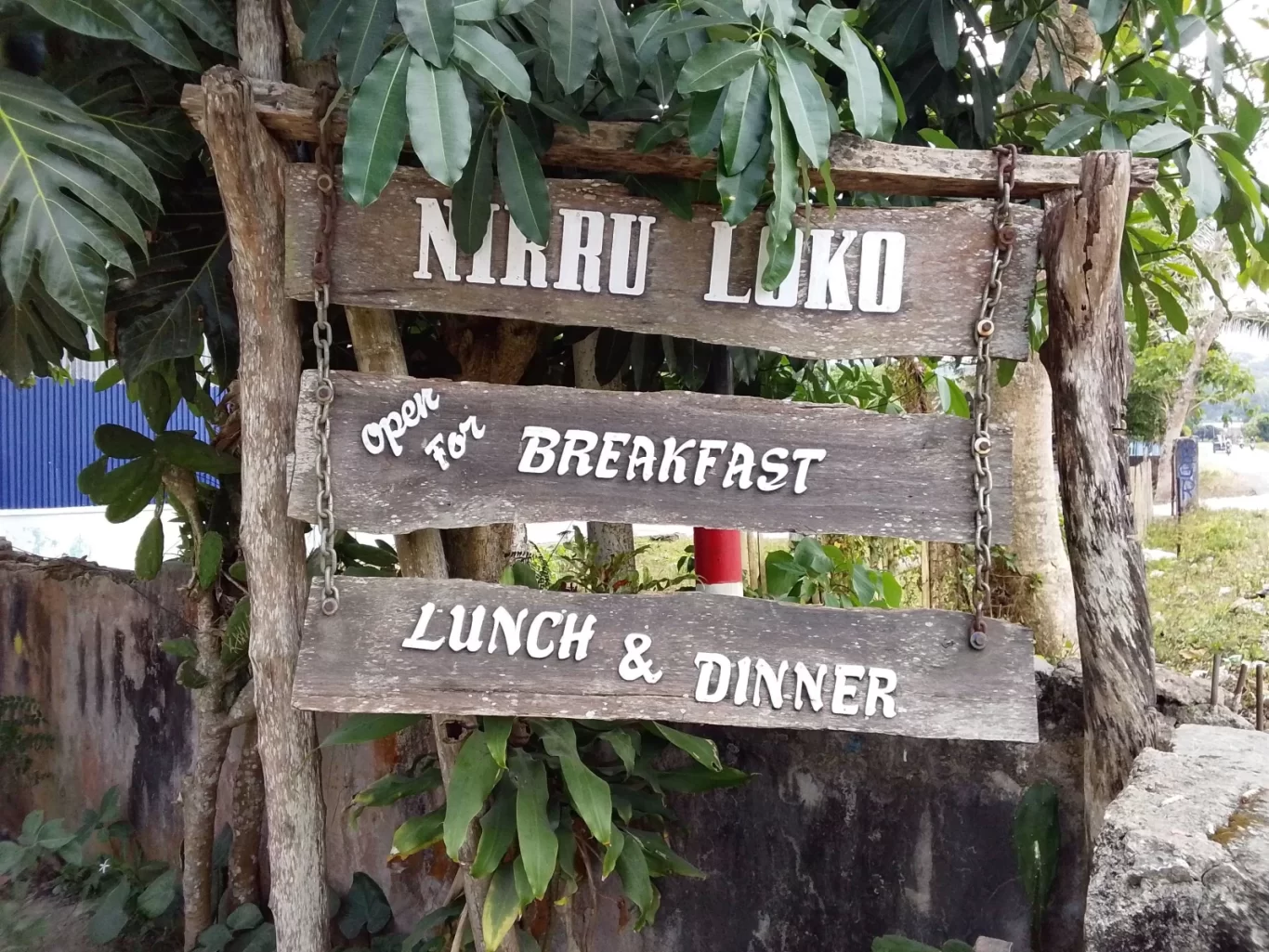 A weathered wooden sign for "Nirru Loko" hangs among green leaves, displaying "Open for Breakfast, Lunch & Dinner" in white text.