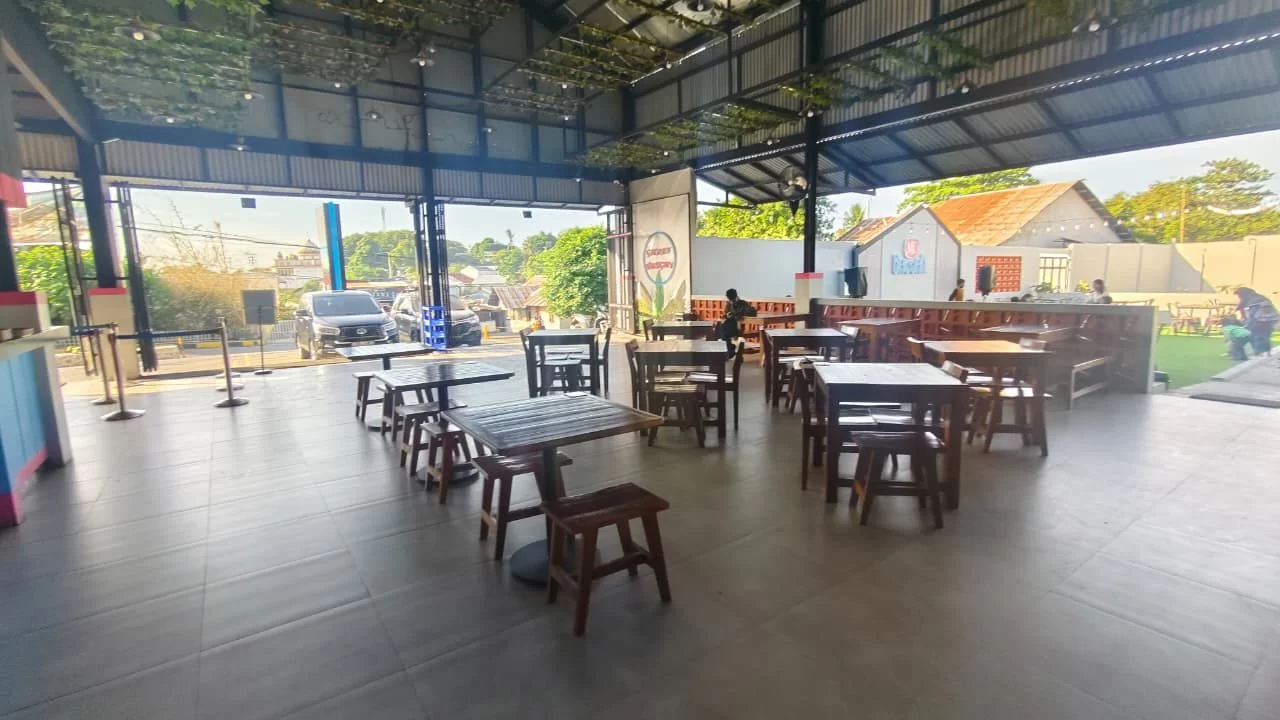 A spacious, open-air cafe features numerous dark wooden tables and stools on a grey tiled floor, with a street and buildings visible outside.