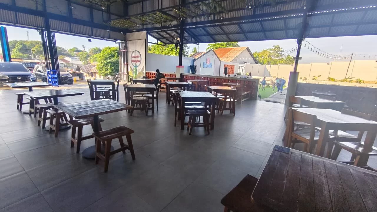 Open-air restaurant with dark wooden tables and stools on gray tiles, overlooking a street with parked cars and distant buildings under a bright sky.