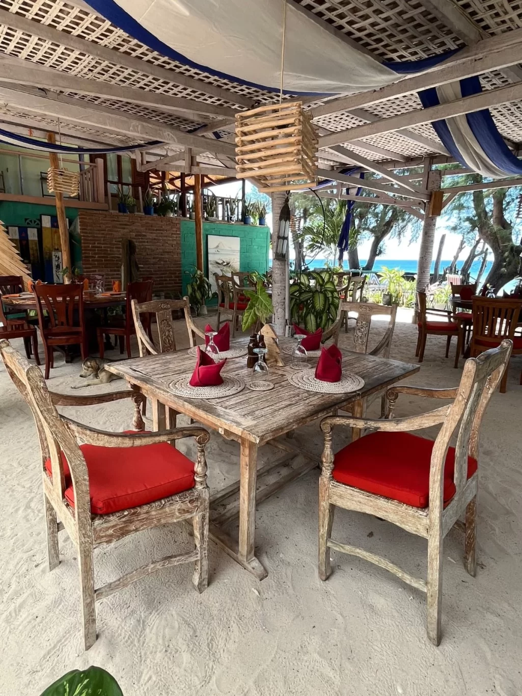 A rustic wooden dining table with red-cushioned chairs sits on white sand under a draped roof, overlooking the blue ocean.