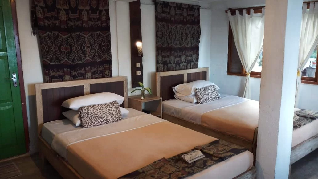 A simple hotel room features two single beds with light brown blankets and white pillows, accented by dark patterned tapestries on white walls. A green door is on the left.