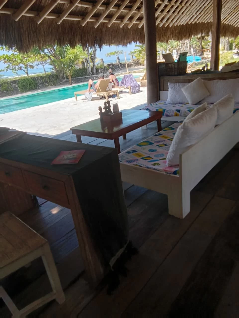 A bright open-air beachfront suite with a thatched roof, wooden furniture, and white daybeds overlooks a turquoise pool and the ocean.