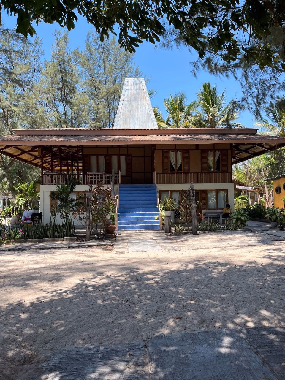 A two-story traditional Sumba-style building with a blue staircase and corrugated metal roof stands on sandy ground, framed by trees under a clear sky.