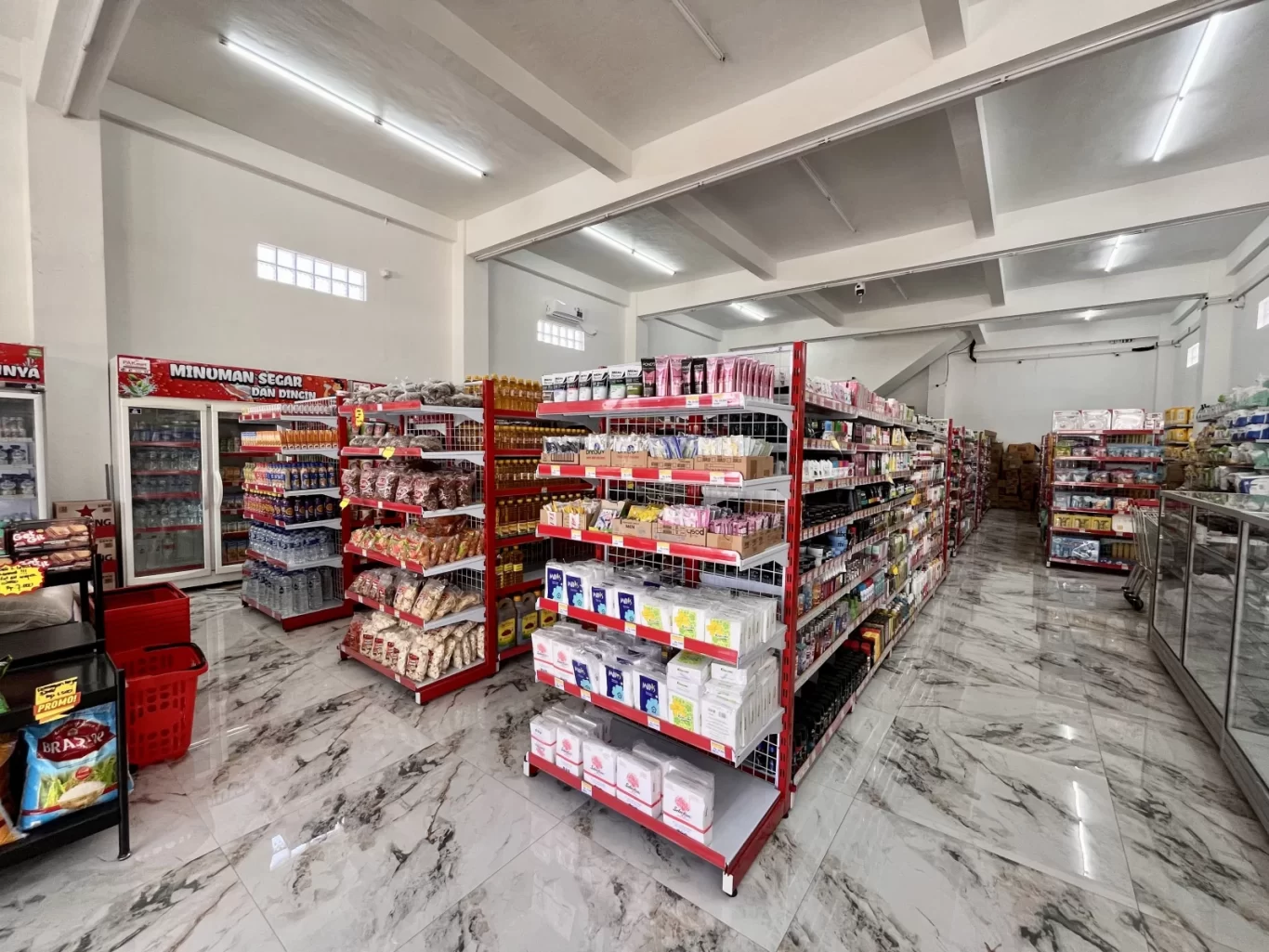 Interior of a bright, modern minimarket with white walls and marble floors. Red and white shelves are stocked with various packaged goods, and refrigerators line the back wall.