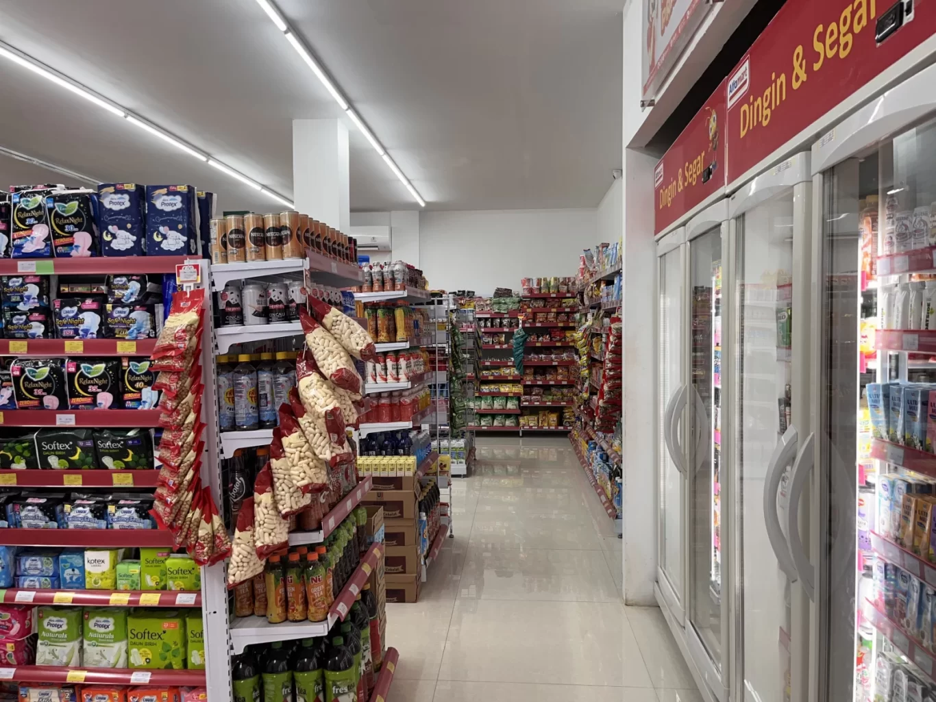 Interior view of a brightly lit minimarket aisle in Loura, Sumba, with shelves stocked with diverse packaged goods and refrigerators on the right.