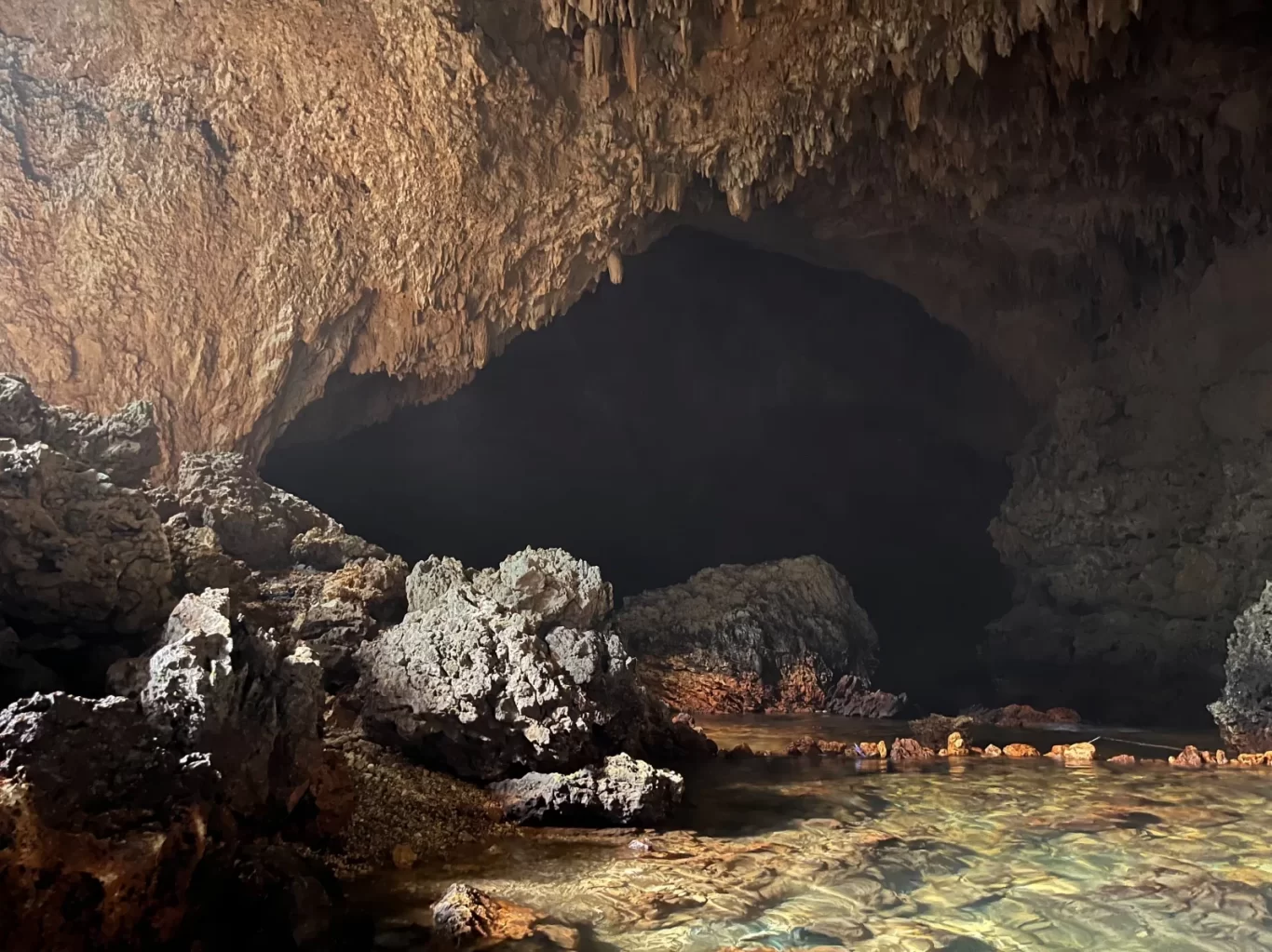 A dark cave interior, Gua Weli'a, features a clear, shallow spring pool reflecting the rocky bottom, surrounded by large brown and gray formations and stalactites.