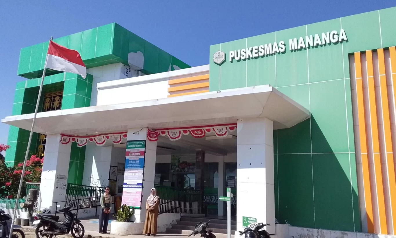 A green and white building with "PUSKESMAS MANANGA" signage, an Indonesian flag, and two people standing near the entrance, under a clear blue sky.