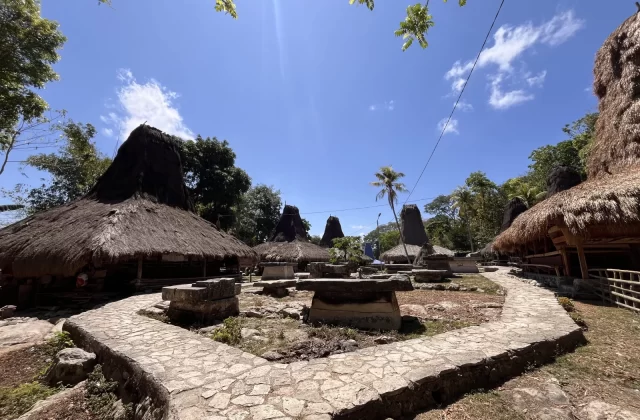 Traditional Sumba village houses with tall thatched roofs and ancient stone megaliths are arranged around a central dirt area with a stone path under a bright blue sky.