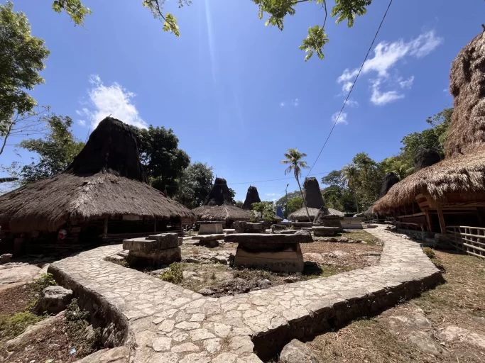 Traditional Sumba village houses with tall thatched roofs and ancient stone megaliths are arranged around a central dirt area with a stone path under a bright blue sky.