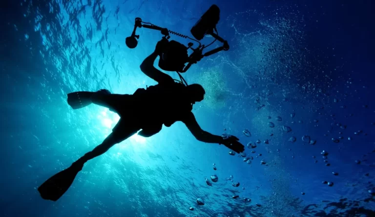 A scuba diver with an underwater camera rig is silhouetted against the bright blue water surface, surrounded by rising bubbles.