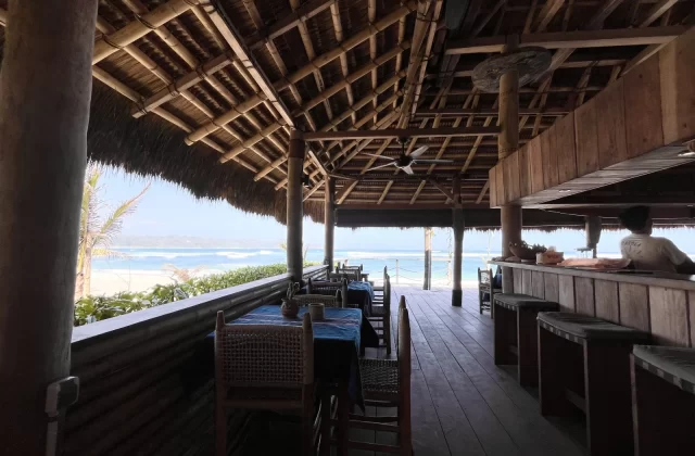 An open-air bar with a thatched roof and wooden interior overlooks a white sand beach, blue ocean, and distant land under a bright sky, with a person behind the bar.