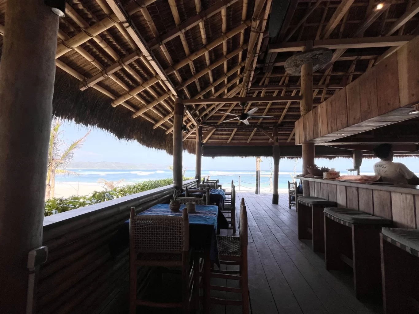 An open-air bar with a thatched roof and wooden interior overlooks a white sand beach, blue ocean, and distant land under a bright sky, with a person behind the bar.