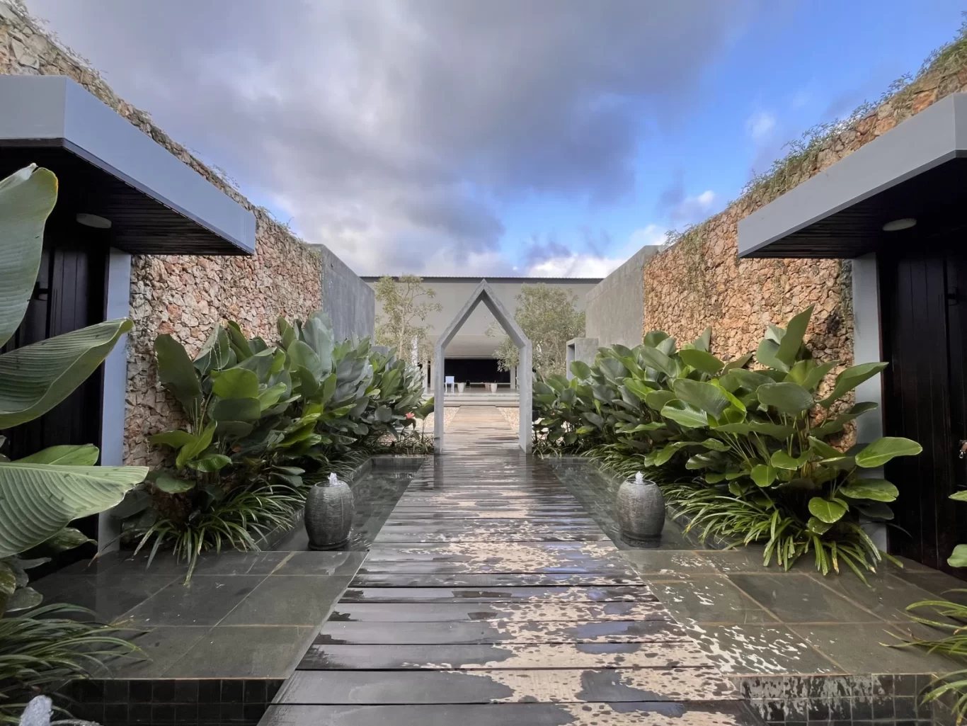 A modern outdoor pathway with dark wood and water features leads to a villa entrance, flanked by lush green plants and stone walls under a cloudy sky.