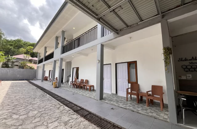 A modern two-story homestay building with white walls, gray pillars, and a metal roof, featuring multiple rooms with outdoor seating. A stone courtyard is in the foreground, with green trees and a cloudy sky in the background.