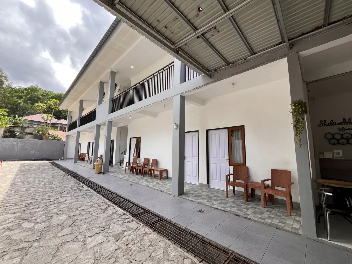 A modern two-story homestay building with white walls, gray pillars, and a metal roof, featuring multiple rooms with outdoor seating. A stone courtyard is in the foreground, with green trees and a cloudy sky in the background.