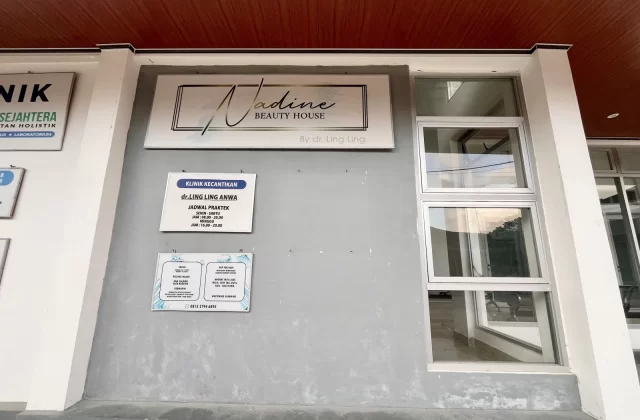 The exterior of Nadine Beauty House clinic in Waikabubak, West Sumba, features a grey wall, large windows, and a brown ceiling, with signs detailing its name and services.