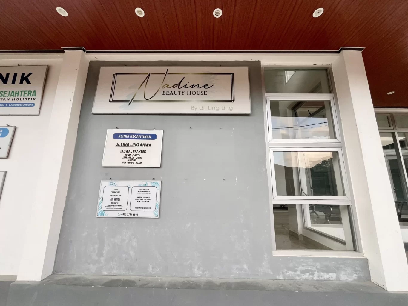 The exterior of Nadine Beauty House clinic in Waikabubak, West Sumba, features a grey wall, large windows, and a brown ceiling, with signs detailing its name and services.