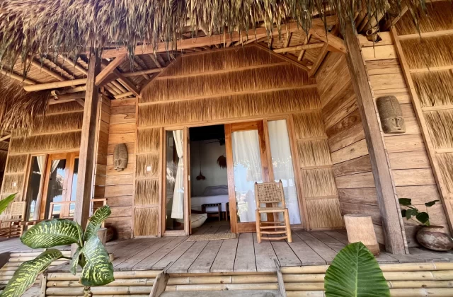 A rustic wooden spa bungalow with a thatched roof and a bamboo porch features an open doorway revealing a bed, surrounded by green tropical plants.